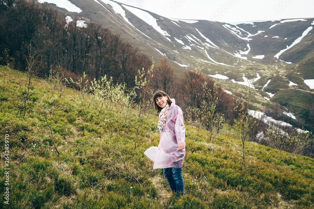 Happy smiling tourist girl in pink rain jacket in mountains surrounded by forest, enjoying silence and harmony of nature