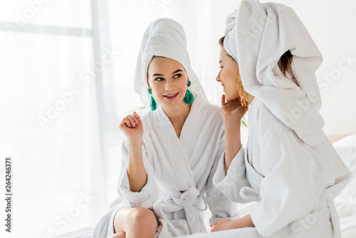 stylish girls in bathrobes, earrings and towels on head talking in bedroom
