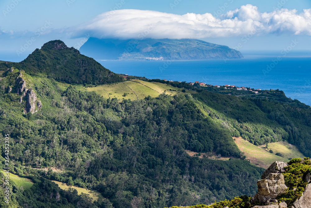 The Azores island of Corvo seen across the ocean from Flores island. There is a wooded landscape in the foreground, with a village. The top of Corvo is hidden in cloud.