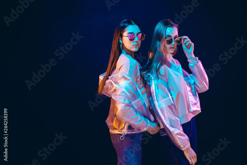 Side view of provocative teen girls in stylish jackets and sunglasses standing on black background in neon illumination