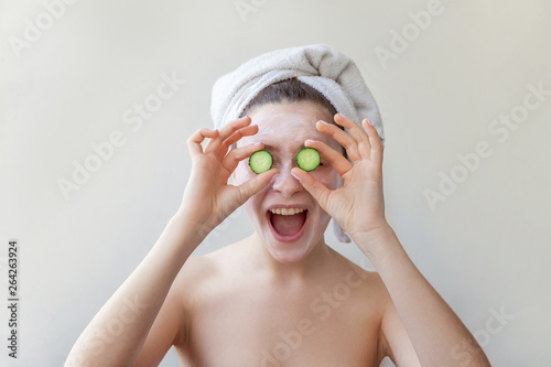 Beauty portrait of woman in towel on head with white nourishing mask or creme on face holding cucumber slices, white background isolated. Skincare cleansing eco organic cosmetic spa relax concept