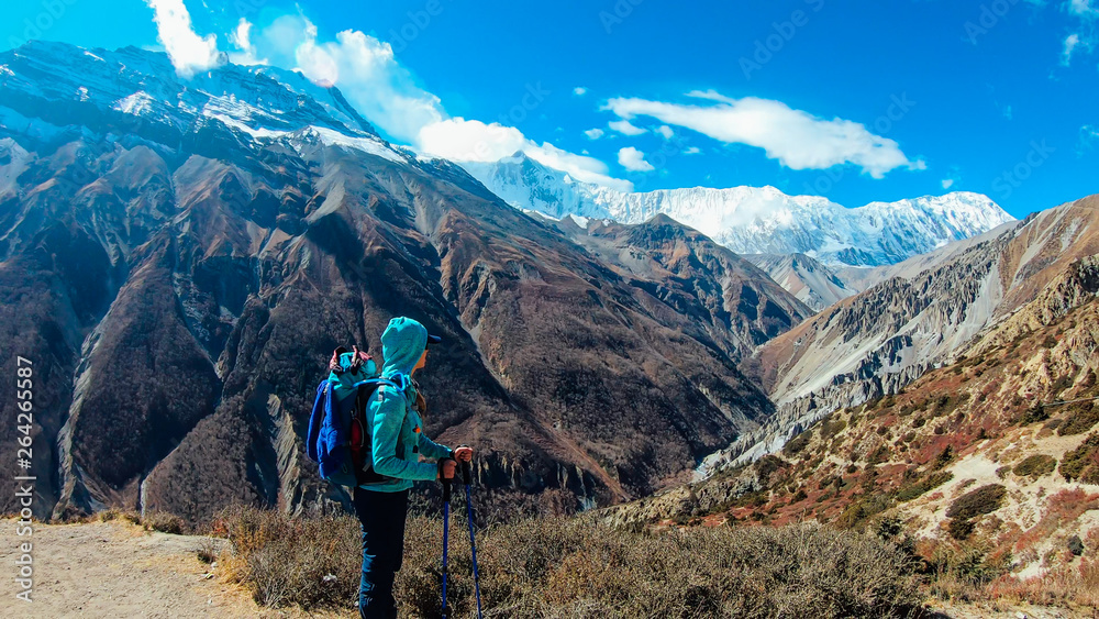 Girl admires Himalayas, carries big backpack, supports herself with sticks, Annapurna Circuit Trek, Nepal. Upper Shreekharka. Dry grass. Socks dry on the backpack. Clouds in the mountains.