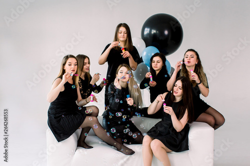 Happy beautiful women celebrating birthday or hen party, sitting on white sofa indoor and blowing soap bubbles. Birthday party, balloons, holiday