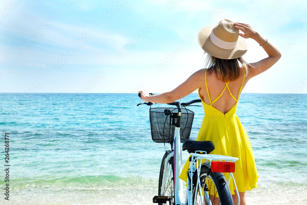 Young carefree woman in bright yellow dress with bicycle at ocean beach. Unrecognizable female wearing broad brim straw hat biking on sandy sea shore on sunny day. Copy space, background. Young carefree woman in bright yellow dress with bicycle at ocean beach. Unrecognizable female wearing broad brim straw hat biking on sandy sea shore on sunny day. Copy space, background.