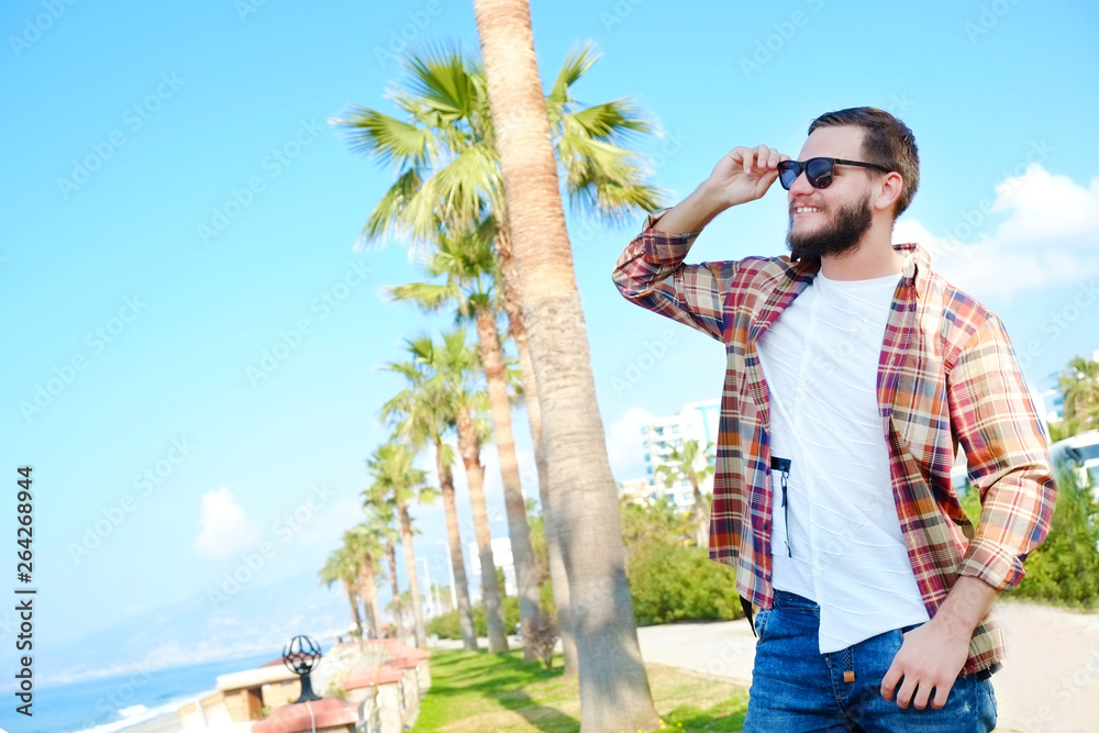 Bearded hipster guy wearing vintage plaid checkered shirt at tropical destination. Portrait of young man in white t-shirt smiling over exotic background. Copy space, close up.