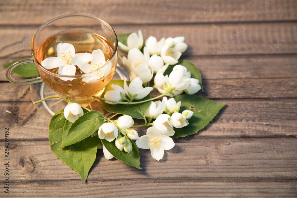 Glass cup with hot fragrant jasmine tea on a wooden background. Around a cup leaflets and flowers of a jasmine are spread out. On a background a green lawn with the blossoming clover Glass cup with hot fragrant jasmine tea on a wooden background. Around a cup leaflets and flowers of a jasmine are spread out. On a background a green lawn with the blossoming clover