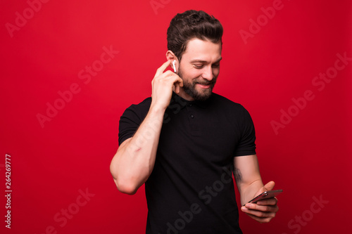 Portrait of happy bearded man singing and listening to music with cell phone and wireless earphones isolated over red wall © F8  \ Suport Ukraine