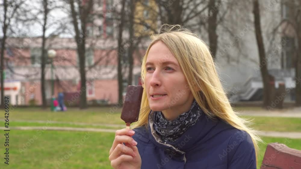 Blonde woman eating ice cream with chocolate icing on a stick. Girl eats ice cream in a park, on a city street. Blonde woman eating ice cream with chocolate icing on a stick. Girl eats ice cream in a park, on a city street.