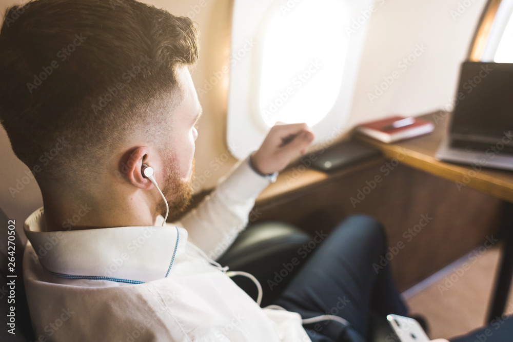 Fototapeta premium Young attractive and successful businessman is resting and listening to music works while sitting in the chair of his private business plane