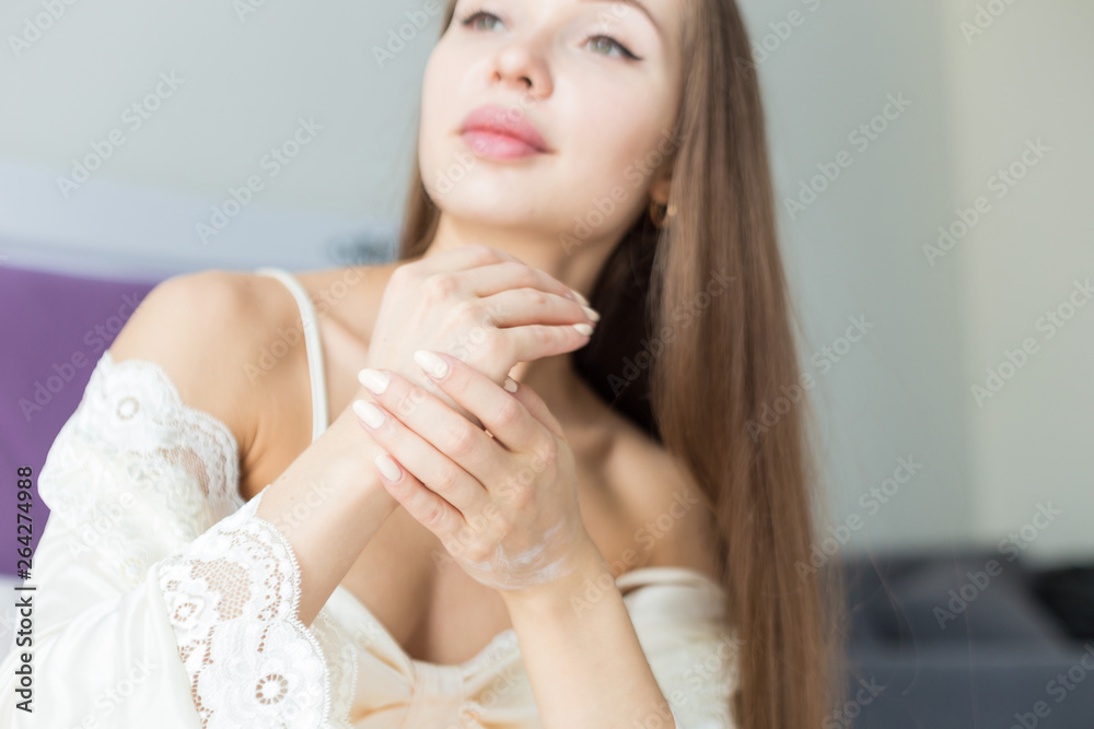 Naklejka premium Attractive young girl in night clothes applies cream to skin and rubs it while doing morning procedures. Moisturizing and skin care