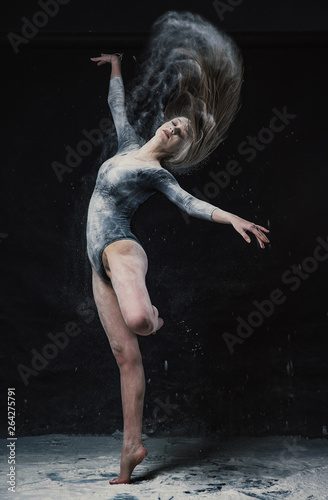 Fototapeta Naklejka Na Ścianę i Meble -  A young beautiful girl in a swimsuit and a translucent skirt emotionally dancing on a black background in clouds of white dust