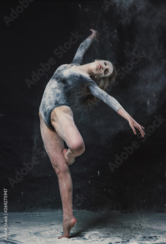 A young beautiful girl in a swimsuit and a translucent skirt emotionally dancing on a black background in clouds of white dust