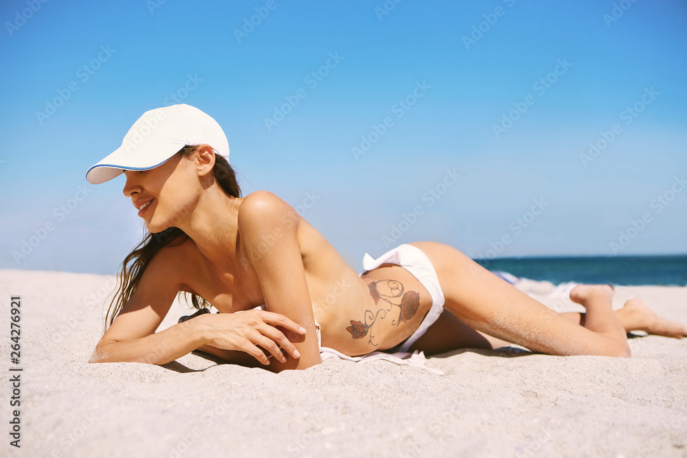 beautiful athletic woman tanning in bikini and white cap on sandy beach at summer. Model with sexy slim body is relaxing lying down on white sand tropical getaway. Summer vacation concept. Side view. beautiful athletic woman tanning in bikini and white cap on sandy beach at summer. Model with sexy slim body is relaxing lying down on white sand tropical getaway. Summer vacation concept. Side view.