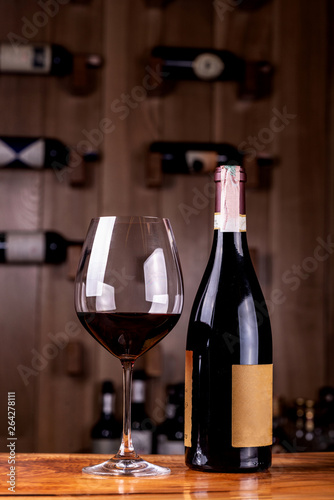 Glass and bottle with delicious red wine on wooden table. In the background Red wine bottles stacked on wooden racks shot with limited depth of field.