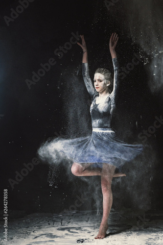 A young beautiful girl in a swimsuit and a translucent skirt emotionally dancing on a black background in clouds of white dust