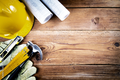 hammer, protective gloves, folding ruler, model knife, blueprint  and yellow safety helmet on wooden background