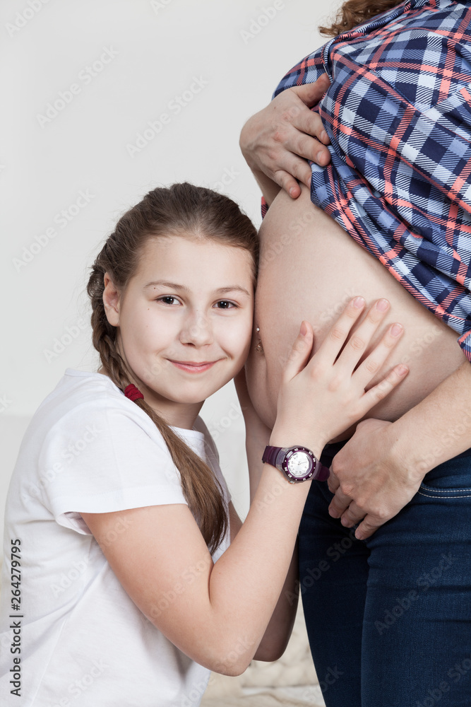 Joyful young girl hugging her pregnant mother tummy, listen and looking at camera and smiling Joyful young girl hugging her pregnant mother tummy, listen and looking at camera and smiling