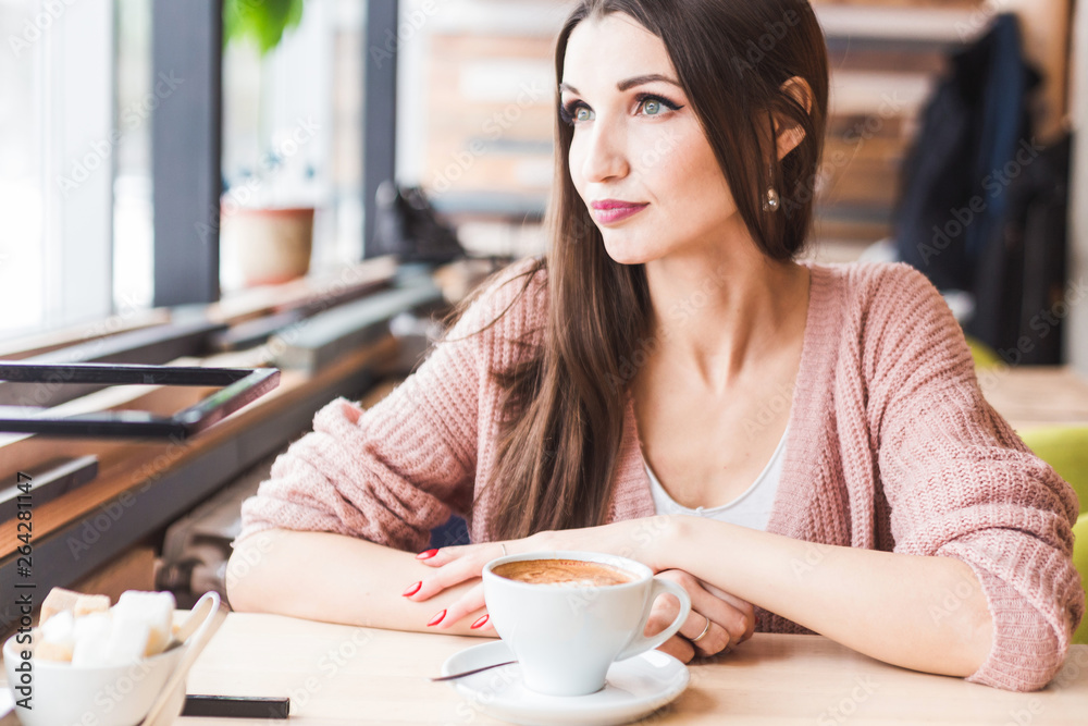 Beautiful young woman sits at a table in a cafe with a cup of coffee and looks out the window Beautiful young woman sits at a table in a cafe with a cup of coffee and looks out the window