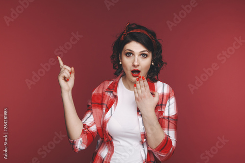 Attractive woman pointing something by her finger on the red background in the studio