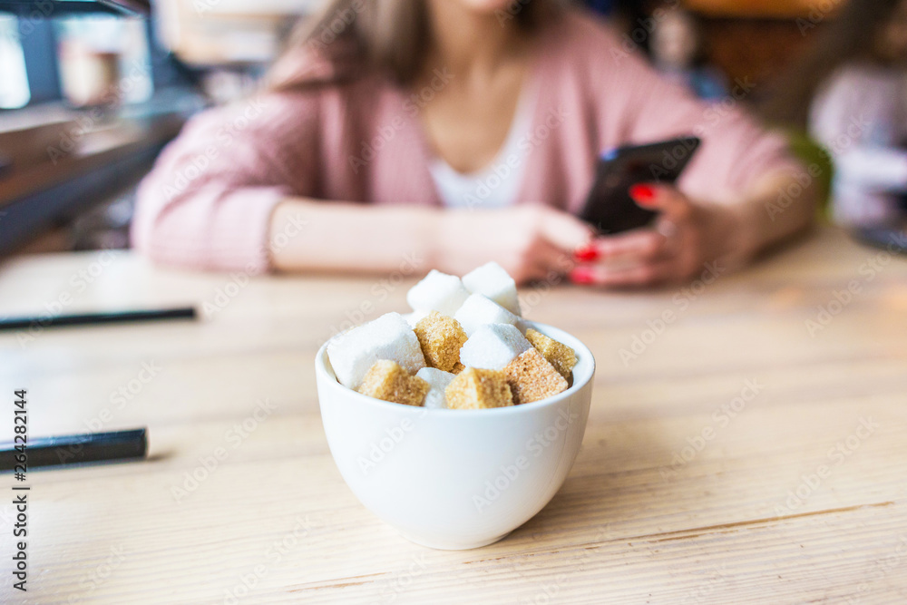 Closeup of a sugar bowl with sugar cubes. In the background, the girl at the table in the interior of the cafe uses the phone