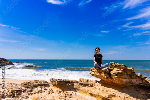 Young woman sitting on cliff on beach in Nazare, Portugal.