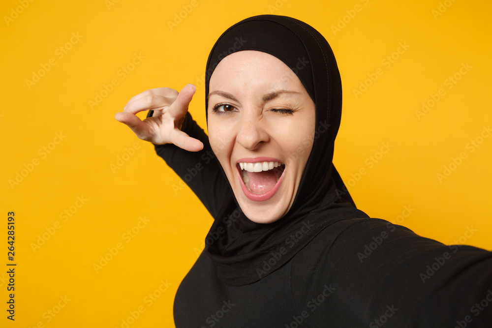Close up young arabian muslim woman in hijab black clothes doing selfie shot on mobile phone isolated on yellow wall background studio portrait. People religious lifestyle concept. Mock up copy space. Close up young arabian muslim woman in hijab black clothes doing selfie shot on mobile phone isolated on yellow wall background studio portrait. People religious lifestyle concept. Mock up copy space.
