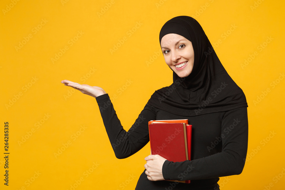 Arabian muslim student girl in hijab black clothes holds books isolated on yellow wall background, studio portrait. People religious lifestyle, education in high school concept. Mock up copy space. Arabian muslim student girl in hijab black clothes holds books isolated on yellow wall background, studio portrait. People religious lifestyle, education in high school concept. Mock up copy space.
