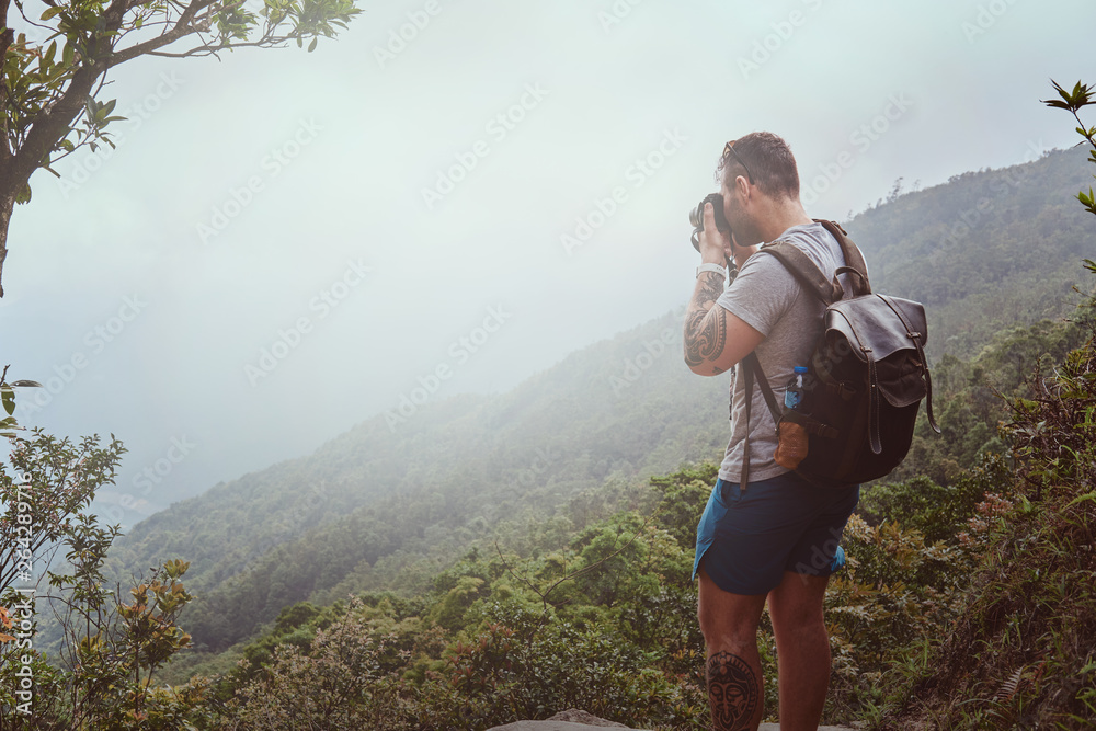 Young man with backpack is taking photo of beautiful nature on his photo camera while hiking in mountains.