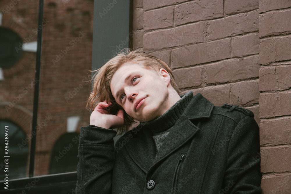 young blond guy standing against the wall and holding a strand of hair, close-up portrait