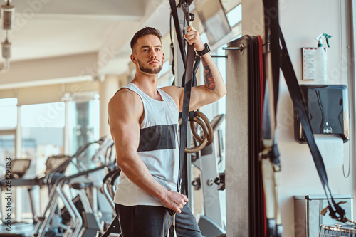 Attractive young bodybuilder is posing next to training apparatus at sunny gym.