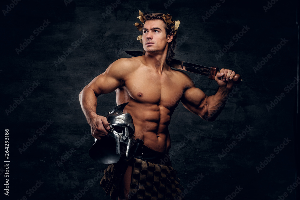 Fototapeta premium Attractive powerful man in wreath is posing on grey background and holds helmet and sword in his hands.