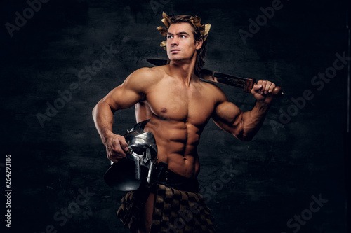 Attractive powerful man in wreath is posing on grey background and holds helmet and sword in his hands.