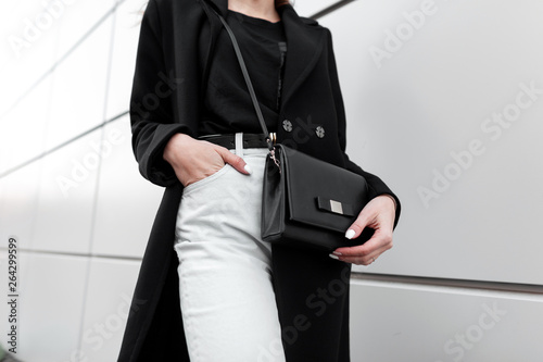 Fotografie Young stylish woman in a fashionable long black coat in vintage white jeans in a black trendy t-shirt with a leather stylish black handbag is standing outdoors