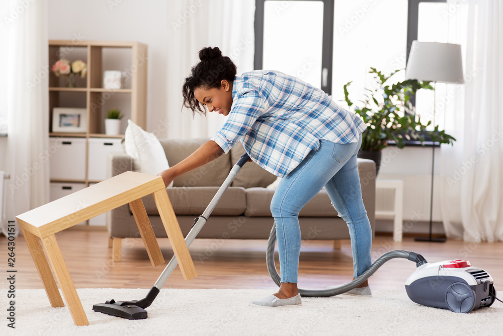 household and housework concept - happy african american woman or housewife with vacuum cleaner cleaning carpet under table at home