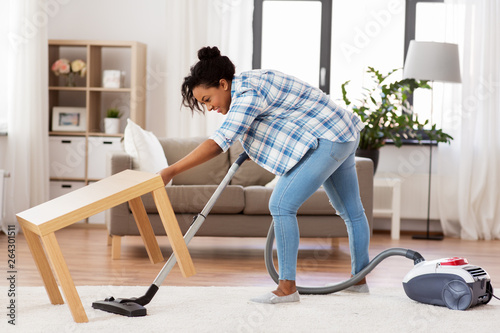 household and housework concept - happy african american woman or housewife with vacuum cleaner cleaning carpet under table at home © Syda Productions