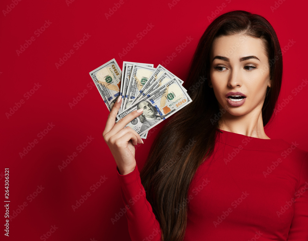 Young brunette woman in tight red dress looks at her hand and is surprised by bundles of dollars notes she holds Young brunette woman in tight red dress looks at her hand and is surprised by bundles of dollars notes she holds