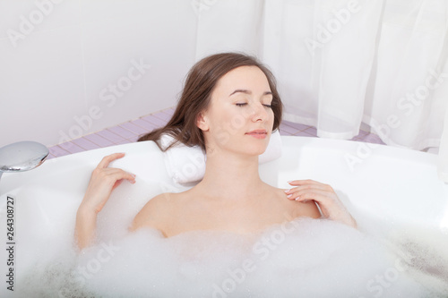 Young pretty woman relaxes in bathtub with soap foam. Spa procedures in bathroom at home or hotelroom. Skin care and moisturizing for youth preservation.