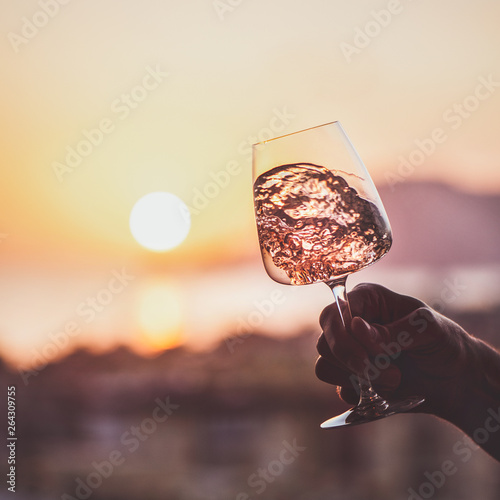 Glass of rose wine in mans hand with sea and sunset at background, close-up, square crop. Summer evening relaxed mood concept