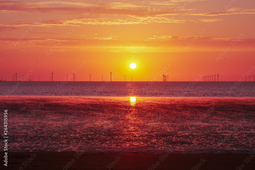 Fototapeta premium Wind turbines and beach at sunset. Reflecting low tide from the vivid sky above. Strong orange, reds and purples. Cloudy sky and the ocean