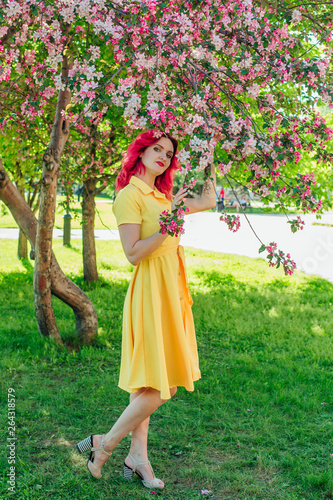 Beautiful and young woman with bright red hair and ref lips standing next to a blooming apple tree in a yellow dress. photo