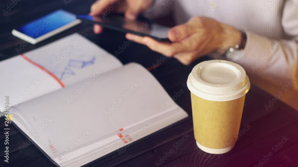 Freelancer at Work Concept. The Businessman in Cafe Сhecks Messages on the Tablet then Checks the Smartphone and Scrolls Through the Notebook. Disposable Paper Cup in the Foreground. Freelancer at Work Concept. The Businessman in Cafe Сhecks Messages on the Tablet then Checks the Smartphone and Scrolls Through the Notebook. Disposable Paper Cup in the Foreground.