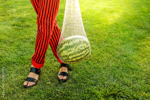 Close up fashion detail, young woman holding net bag with watermelon on a sunny summer day. Food fashion