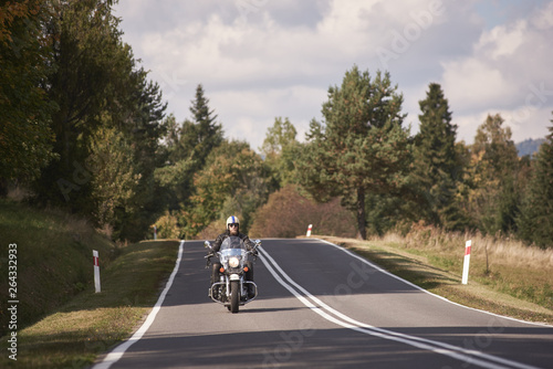 Handsome motorcyclist in black leather outfit riding cruiser bike along beautiful road on bright sunny summer day