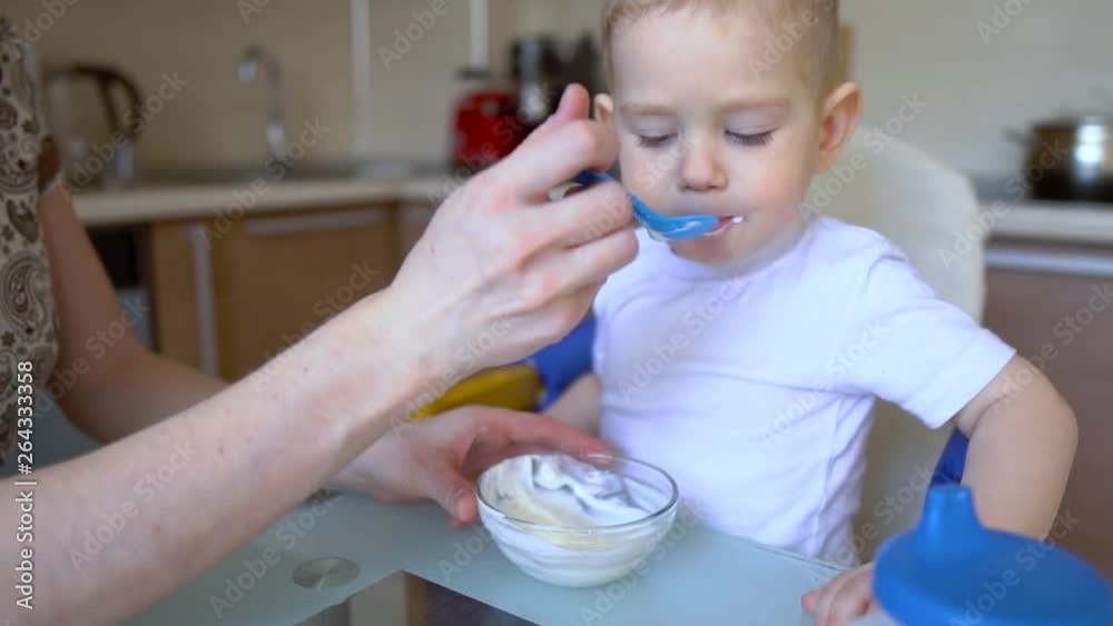 Mother feeding the baby holding out her hand with a spoon of food in the kitchen. Emotions of a child while eating healthy food in slowmotion. Mother feeding the baby holding out her hand with a spoon of food in the kitchen. Emotions of a child while eating healthy food in slowmotion.