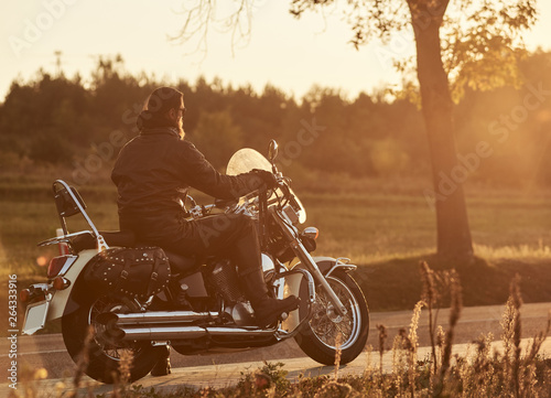 Back view of bearded biker riding cruiser motorcycle along empty narrow country road at sunset on beautifull golden autumn evening landscape background.