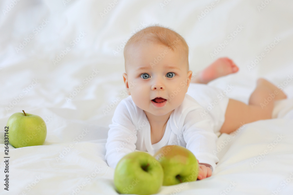 Beautiful blue-eyed child 8 months in white clothes on the bed with green apples Beautiful blue-eyed child 8 months in white clothes on the bed with green apples