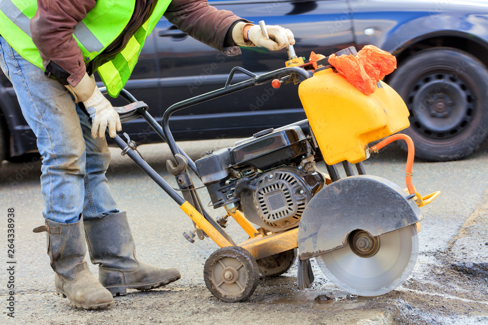 Fototapeta premium A worker in a green reflective vest cuts a piece of bad asphalt with a gasoline cutter on the carriageway.