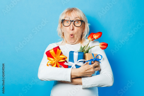 Canvas Print Old woman with a surprised and admiring facial expression holds gifts and a bouquet of tulips in her hands against a blue background