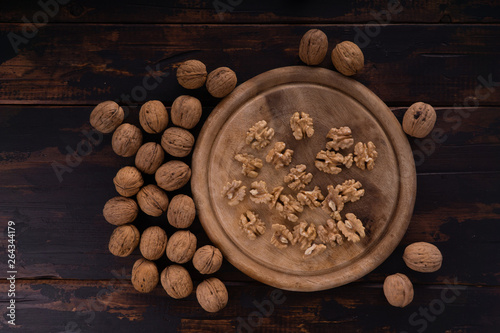 Cracked and whole walnuts lying on round wooden plate and wooden table, top view. Healthy nuts and seeds composition.