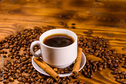 Cup of hot coffee, cinnamon sticks and scattered coffee beans on wooden table